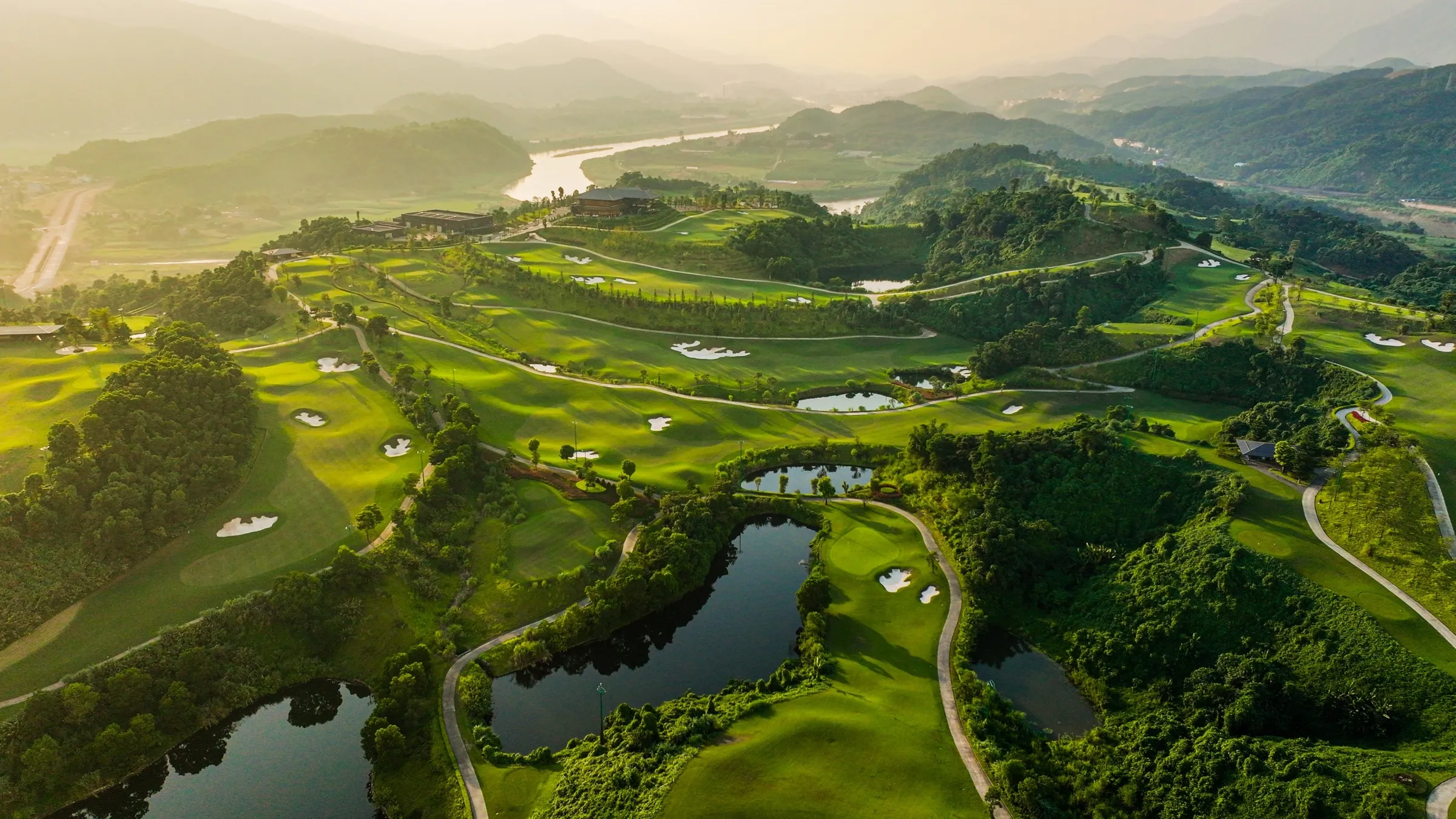 Aerial view of a hillside golf course with lakes, a river and misty valleys at sunrise