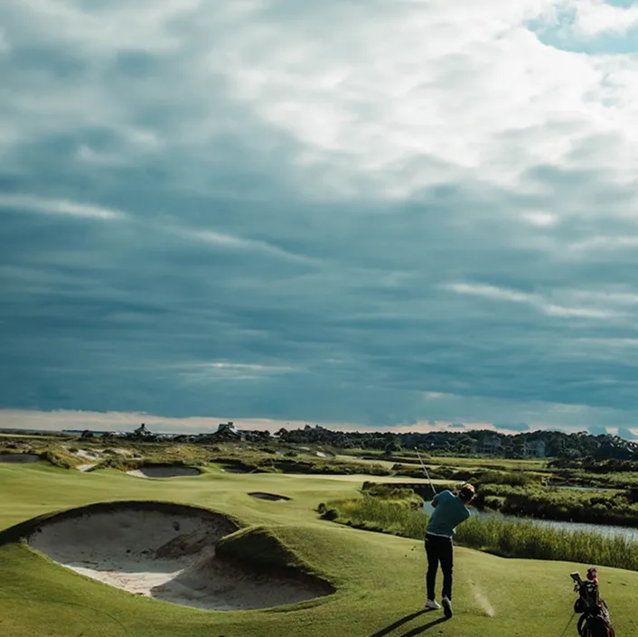 Golfer teeing off beside a sand bunker under a cloudy sky