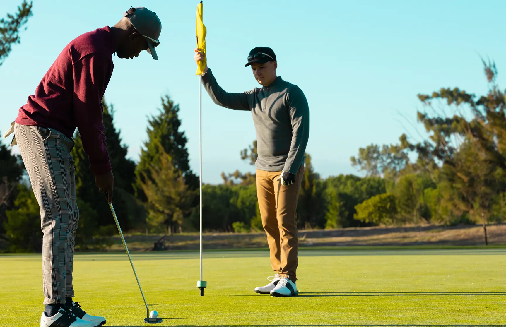 Two golfers on a putting green, one lining up a putt while the other holds the flag