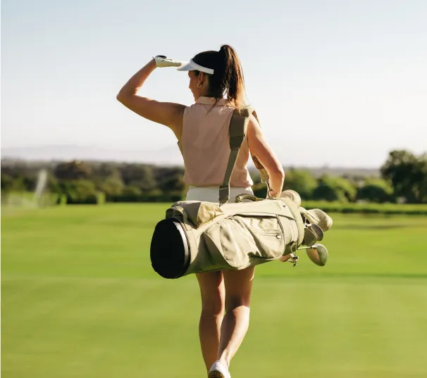 Golfer carrying her bag across a fairway, shading her eyes